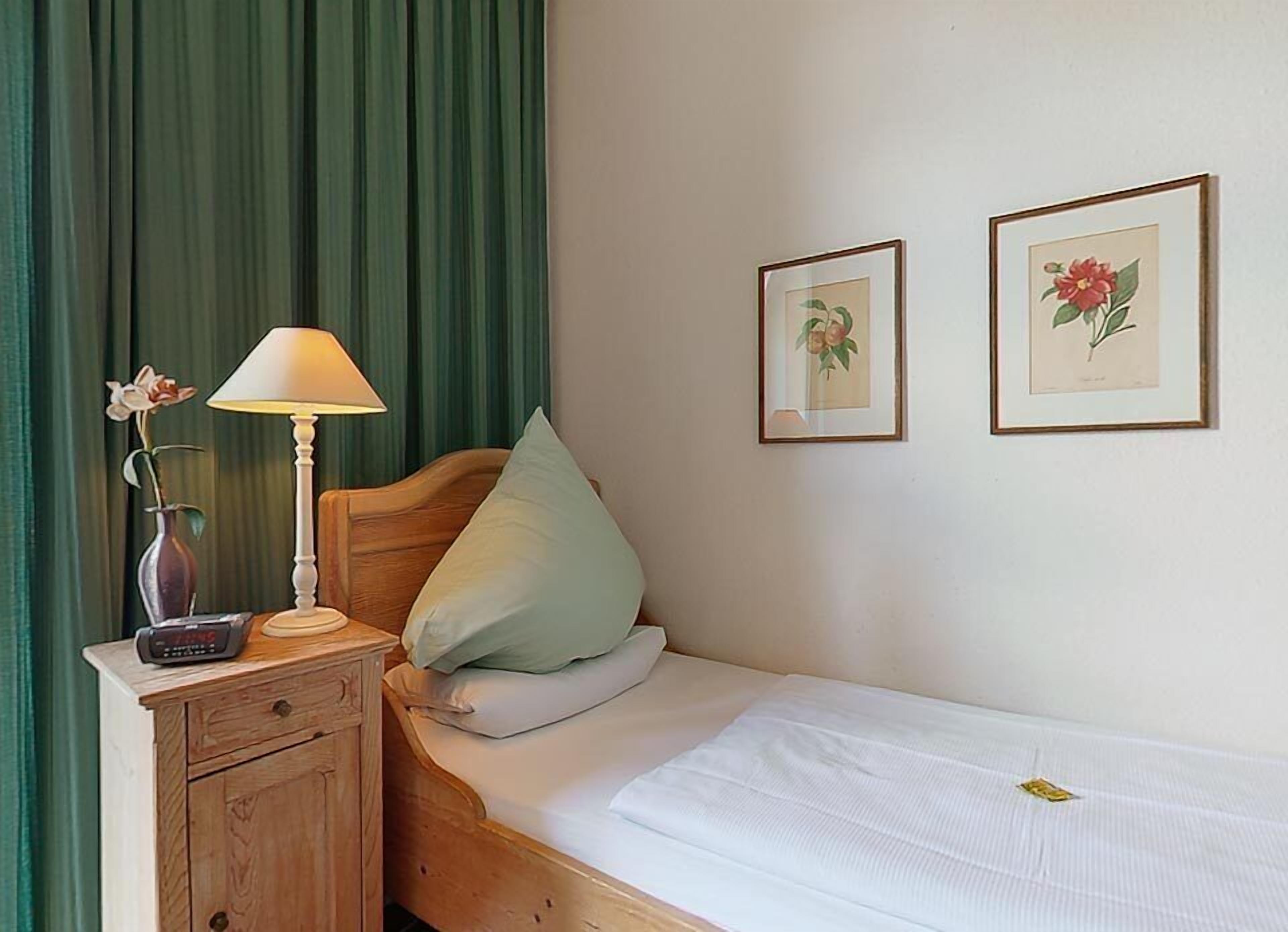 Una cama bien hecha con un almohadón, una mesita de noche con una lámpara, un florero con una flor, y dos láminas botánicas enmarcadas en la pared.