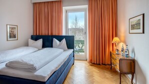 Una cama doble con ropa de cama blanca, un aparador de madera con una lámpara y vistas a un balcón con cortinas naranjas.