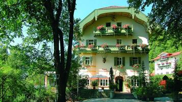Un hotel de estilo arquitectónico europeo tradicional, con una entrada central, varios balcones con jardineras y un jardín bien cuidado.