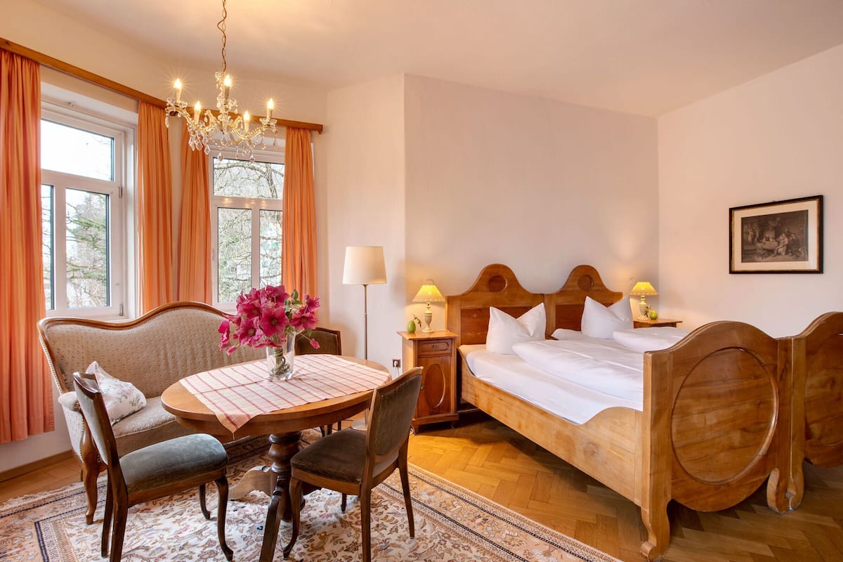 Ein Schlafzimmer mit einem hölzernen Bett, einem Esstisch mit Stühlen, einer Chandelieber und einem Fenster mit Vorhängen.