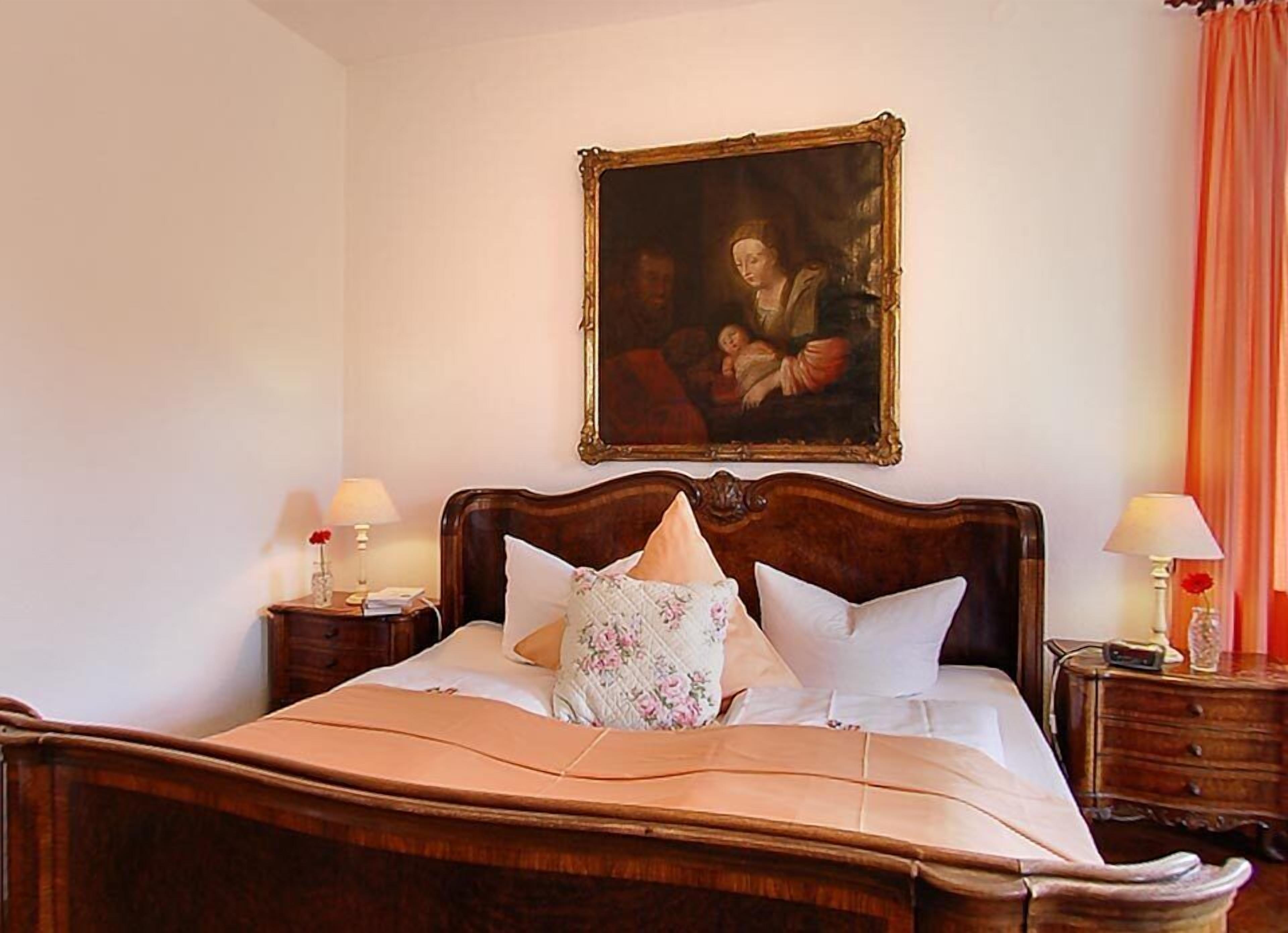 A bedroom with a wooden bed, two bedside tables with lamps, a framed painting above the bed, and a window with curtains.
