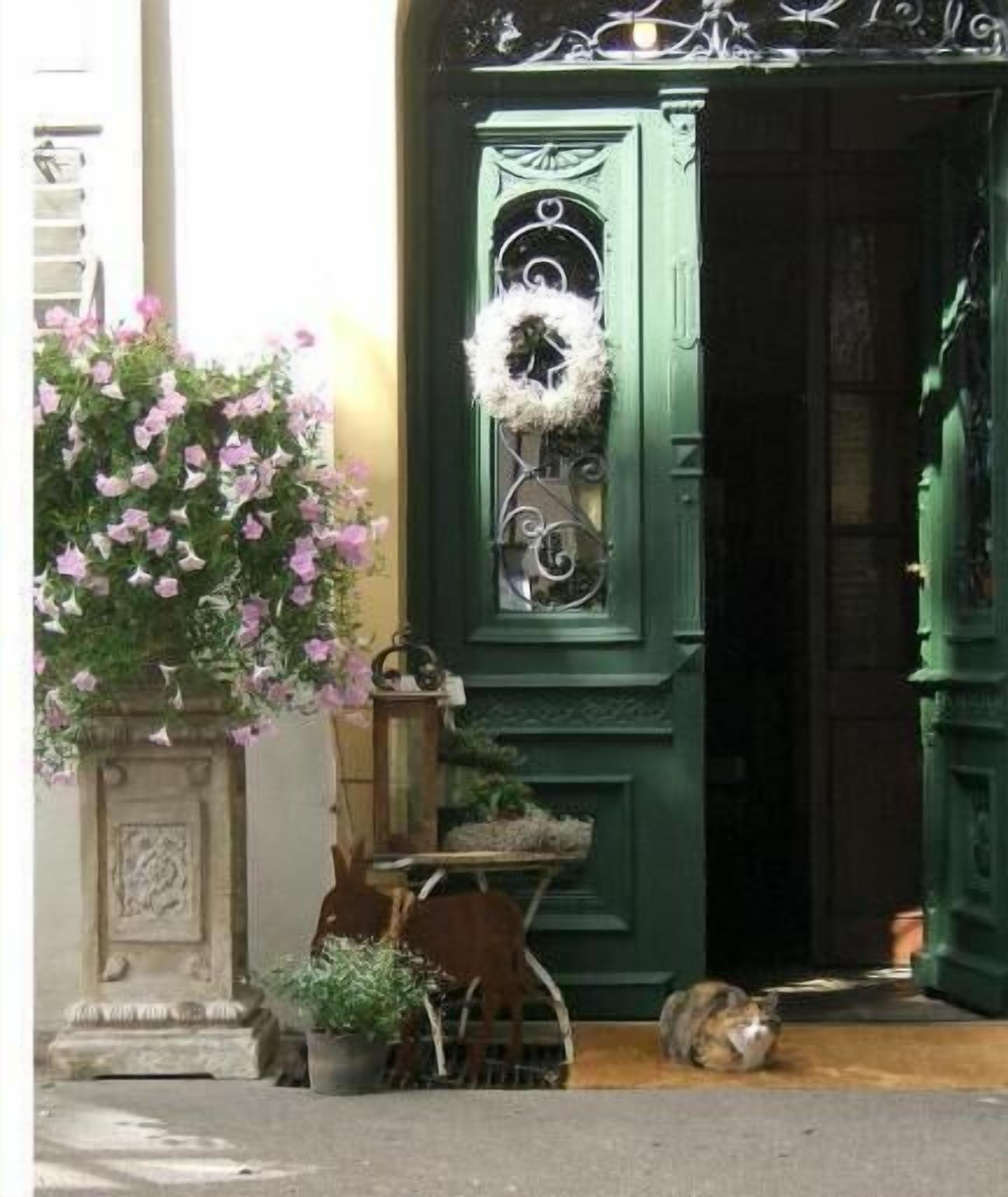Una puerta verde con un adorno de corona, una planta en maceta y una pequeña mesa con una silla frente a un edificio.