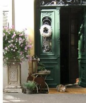 Una puerta verde con un adorno de corona, una planta en maceta y una pequeña mesa con una silla frente a un edificio.