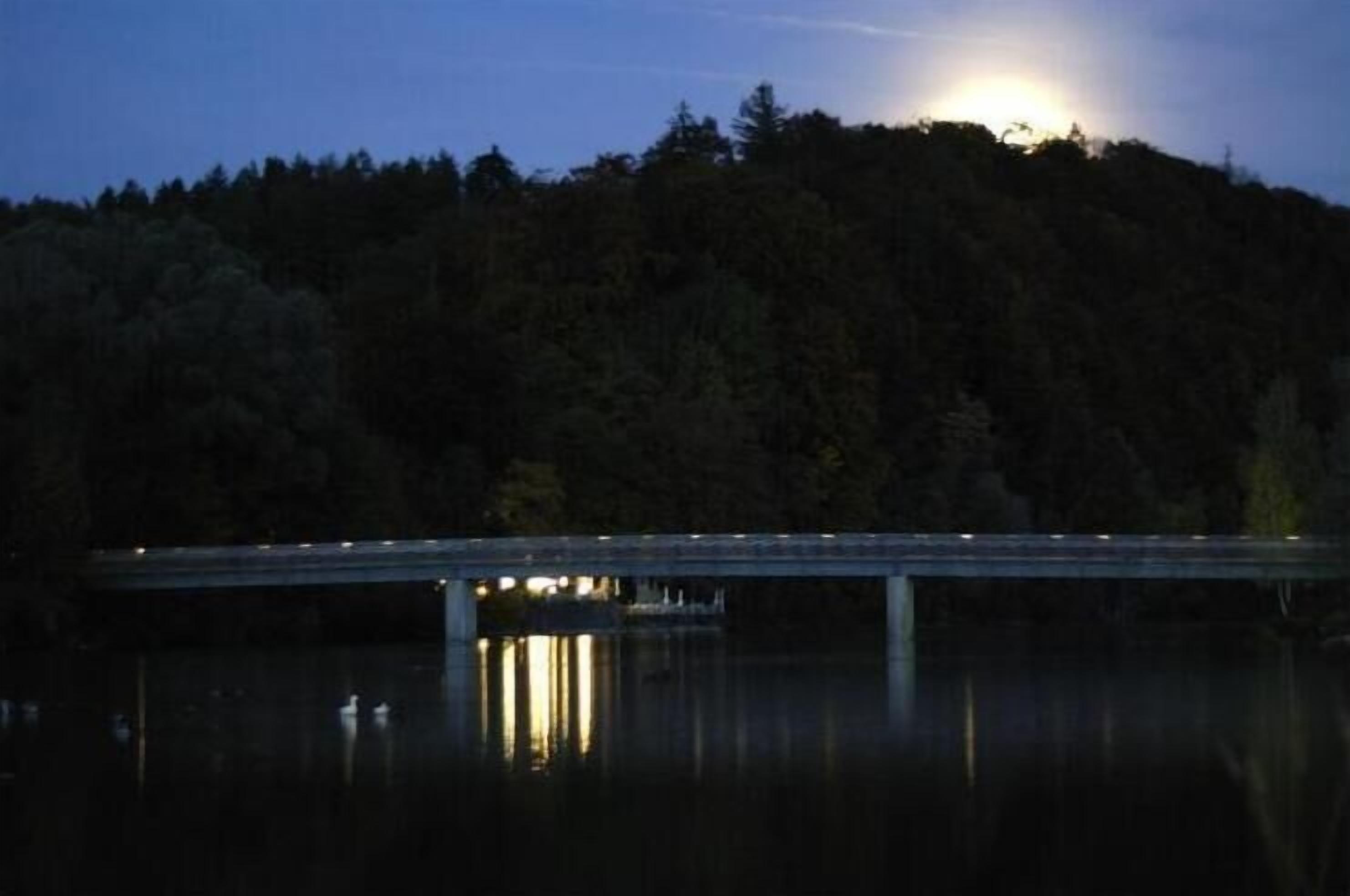 A bridge over a calm body of water with a forested hill in the background.