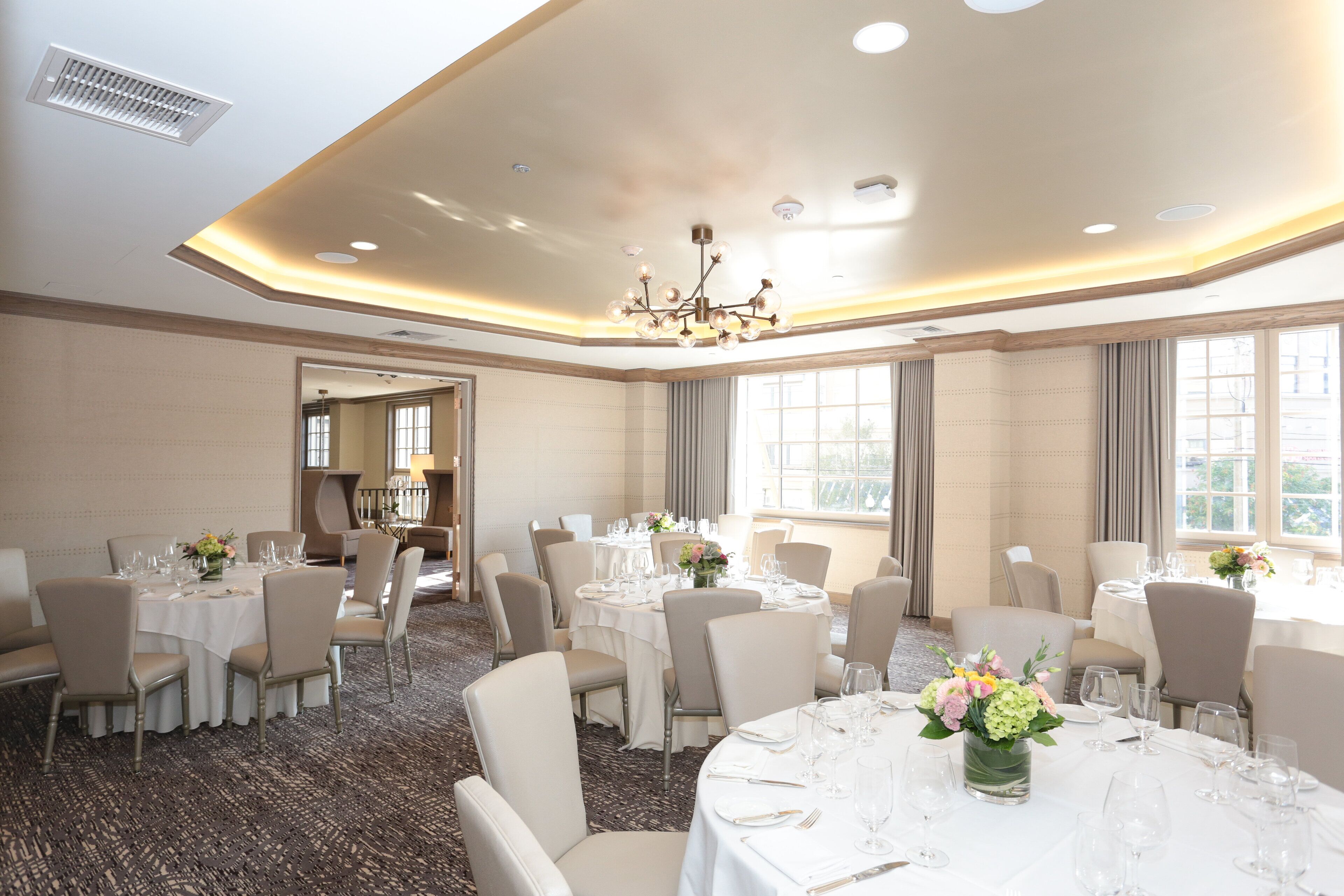 A spacious banquet hall with round tables set for dining, large windows providing natural light, and a modern chandelier.