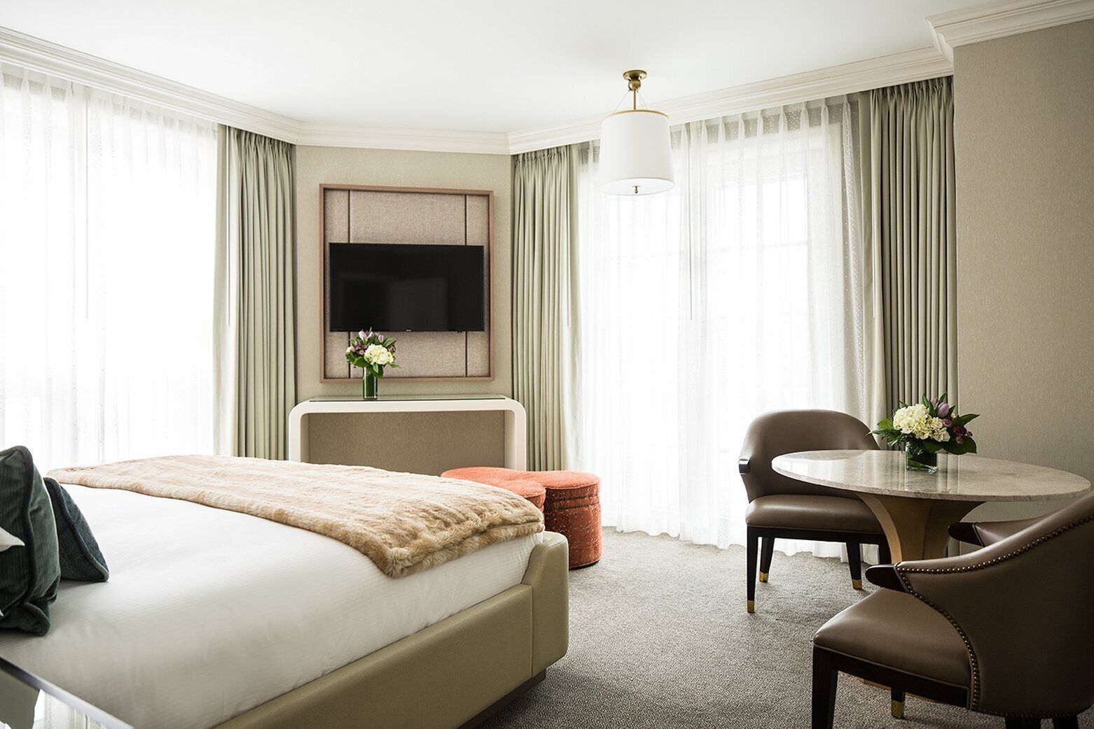 Ein Hotelzimmer mit Bett, Schreibtisch, Stuhl, rundem Tisch, Fernseher und einer Vase mit Blumen.