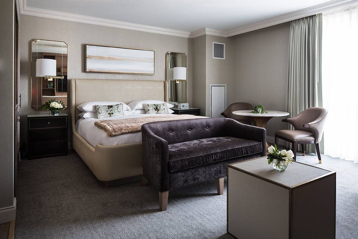 A hotel room with a bed, a dark armchair, a small table, and a chair.