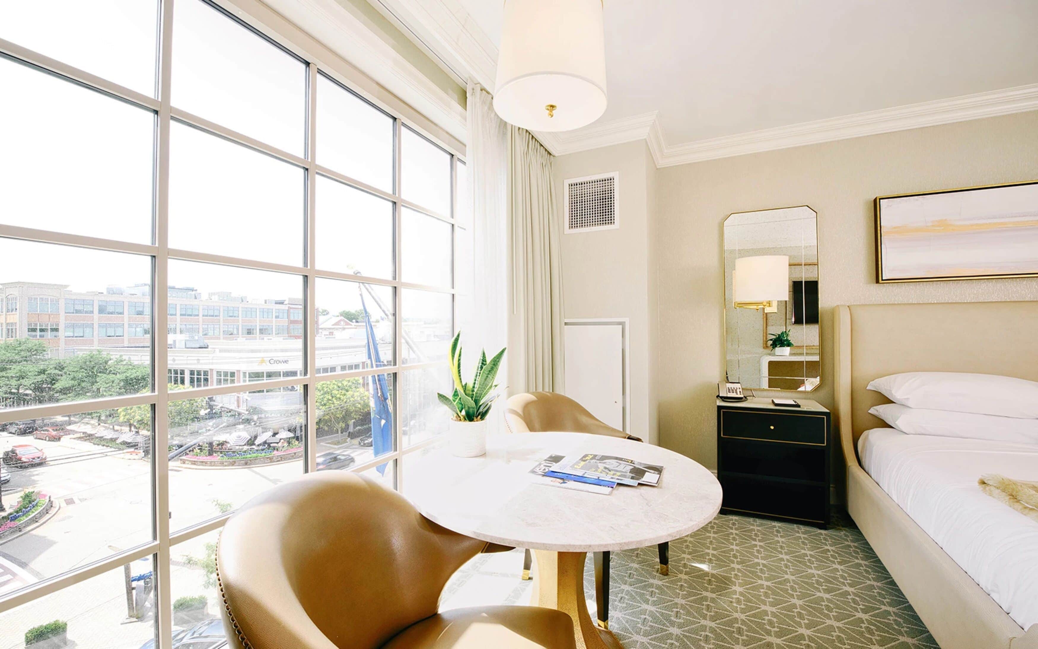 A hotel room with a large window, a round table with a chair, a bed, a bedside table with a lamp, and a mirror on the wall.