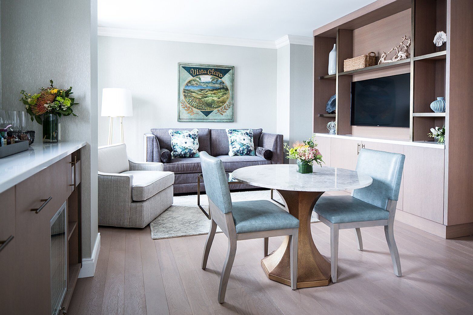 A modern living room with a grey sofa, a round table, and chairs. There is a painting on the wall and a shelf with decorative items.