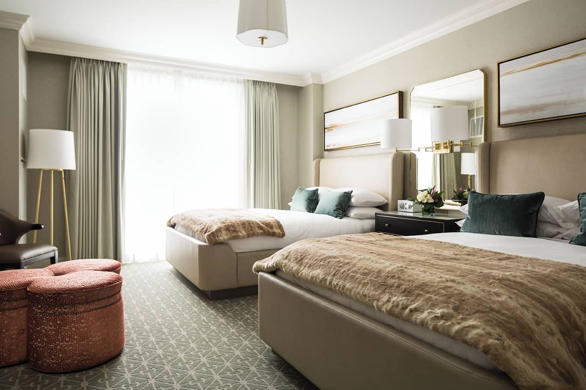 A hotel room with two beds, a sitting area with ottomans, a desk with a lamp, and a large window with curtains.