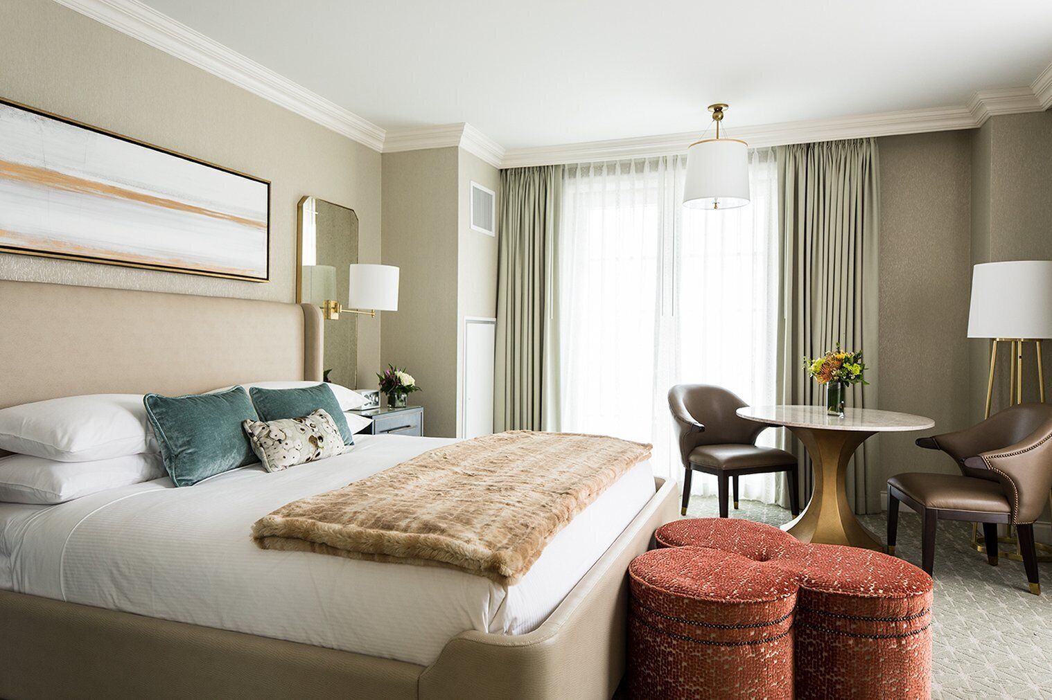 Une chambre d’hôtel avec un grand lit, deux tables de chevet, une table ronde avec des chaises et un petit pouf rouge.