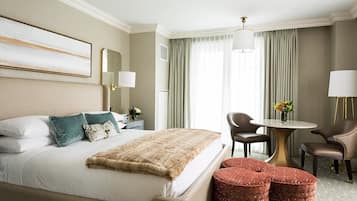 A hotel room with a large bed, two side tables, a round table with chairs, and a small red ottoman.