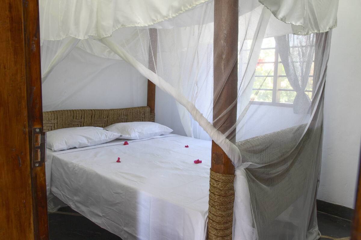 Una cama con sábanas blancas y un mosquitero, cabecera de madera y ventana con vistas a la vegetación.