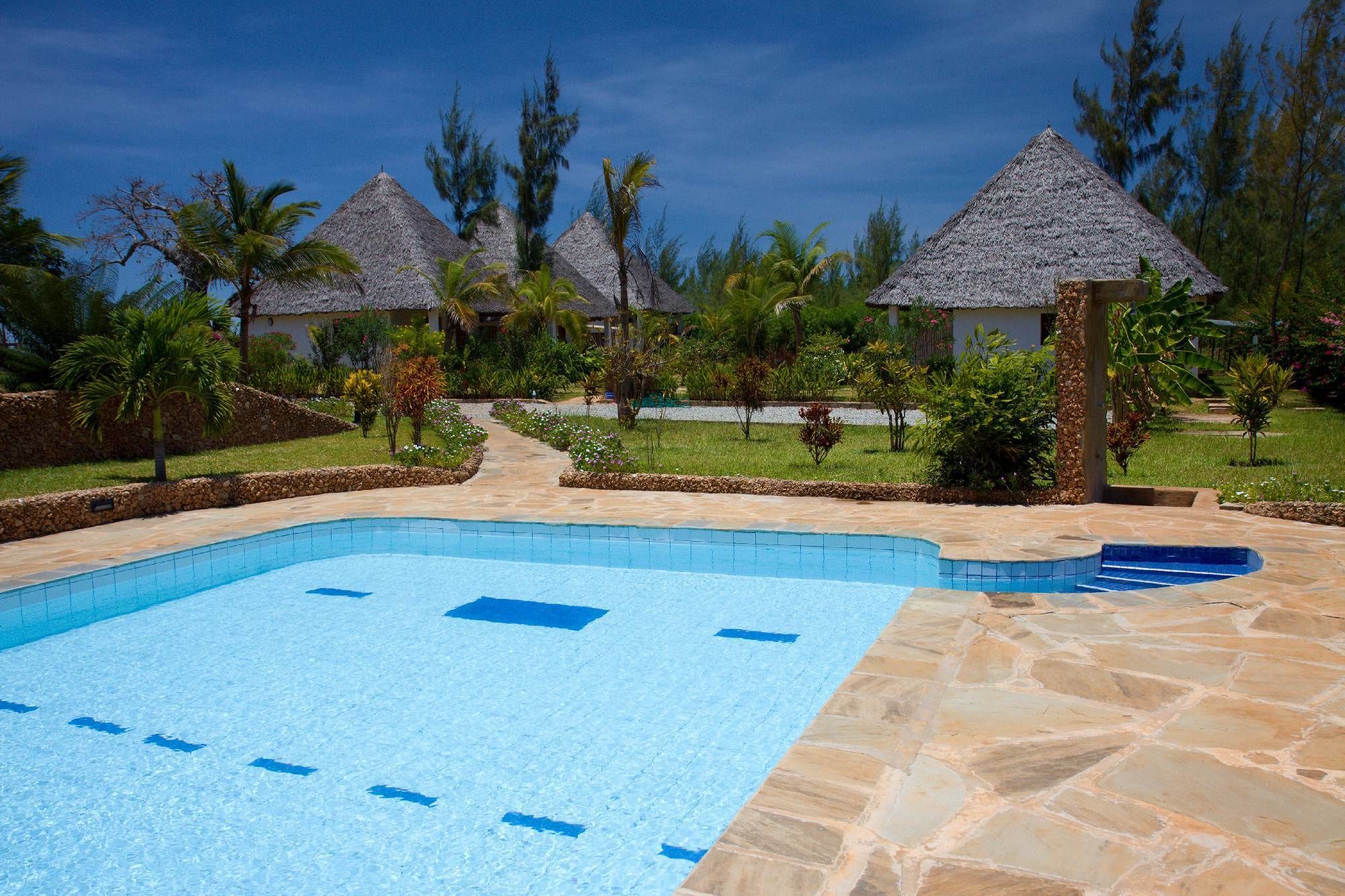 A swimming pool with a stone deck, surrounded by tropical vegetation and thatched-roof structures.