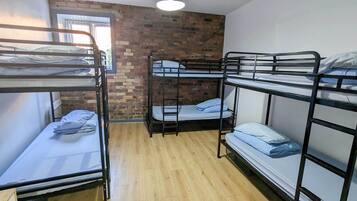 Un dormitorio con literas, suelo de madera, pared de ladrillo y ventana con persianas.