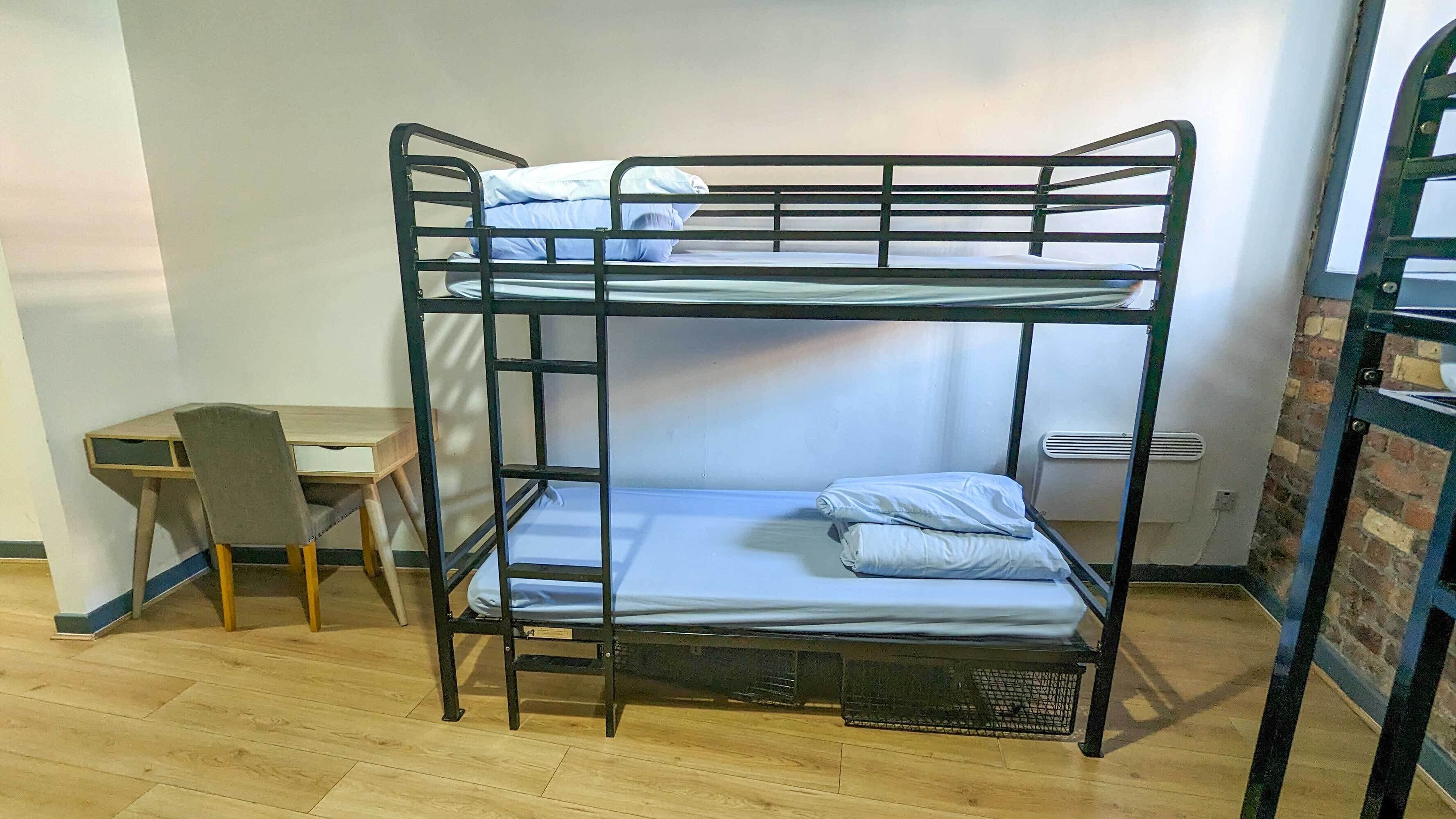 A bunk bed with two levels, a desk, and a chair in a room with wooden flooring and a brick wall.