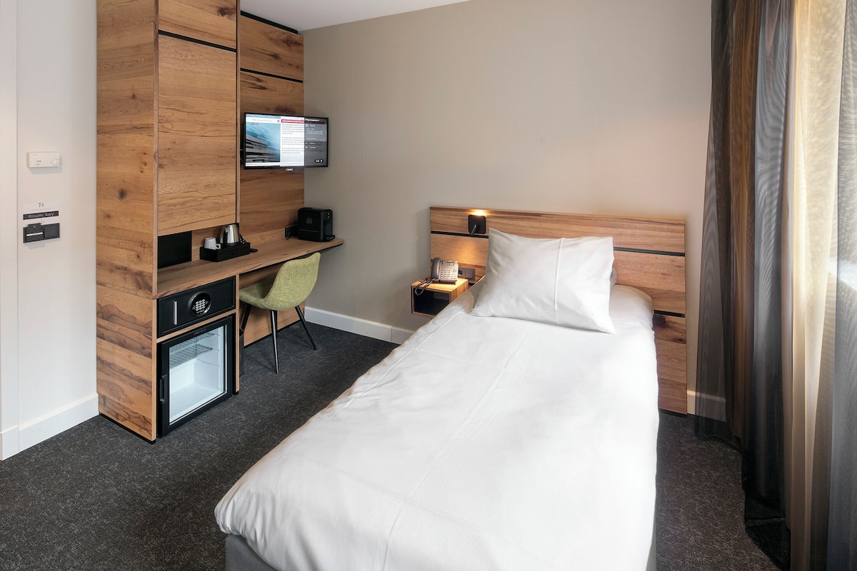 A hotel room with a bed, a desk with a computer, a chair, a small refrigerator, and a television mounted on the wall.