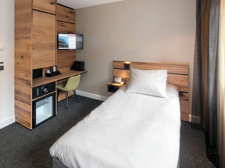 A hotel room with a bed, a desk with a computer, a chair, a small refrigerator, and a television mounted on the wall.