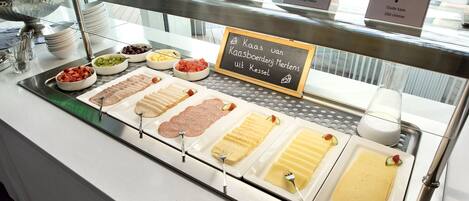 Un bufé con diversos quesos y embutidos, incluyendo queso Gouda y queso Joven, expuestos en un mostrador de vidrio con etiquetas.