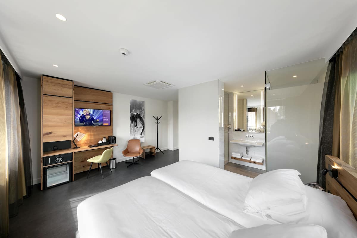A modern hotel room with a large bed, a desk with a TV, a chair, and a glass-enclosed bathroom.