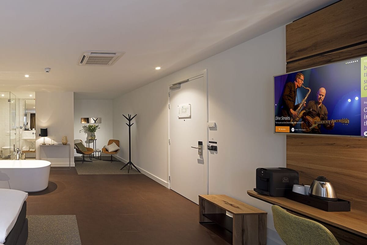 A modern hotel room with a large bed, a bathroom with a bathtub, a TV, and a desk with a kettle.