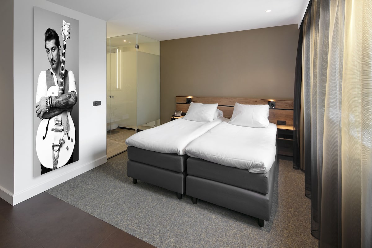 A hotel room with a double bed, a bedside table, a nightstand, a wall-mounted TV, and a poster of a guitar player.