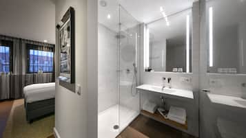Una habitación de hotel moderna con cama, ducha envuelta en vidrio, baño con lavamanos y espejo, y vistas a edificios a través de la ventana.