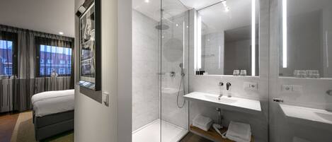 Une chambre d’hôtel moderne équipée d’un lit, d’une douche à parois de verre, d’une salle de bain avec un lavabo et un miroir, et offrant une vue sur les bâtiments par la fenêtre.