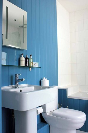 A bathroom with a white sink, a mirror, and a toilet. There are shelves with toiletries. - Montpellier Townhouse (Cheltenham)