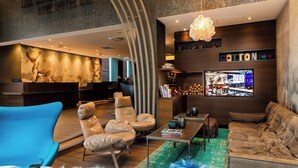 A modern hotel lobby with leather sofas, a blue armchair, a coffee table, and a TV screen displaying a cityscape.
