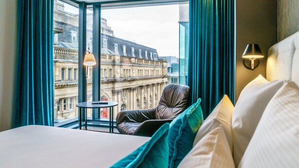 A hotel room with a large window, a leather chair, a small table, and a bed with pillows.