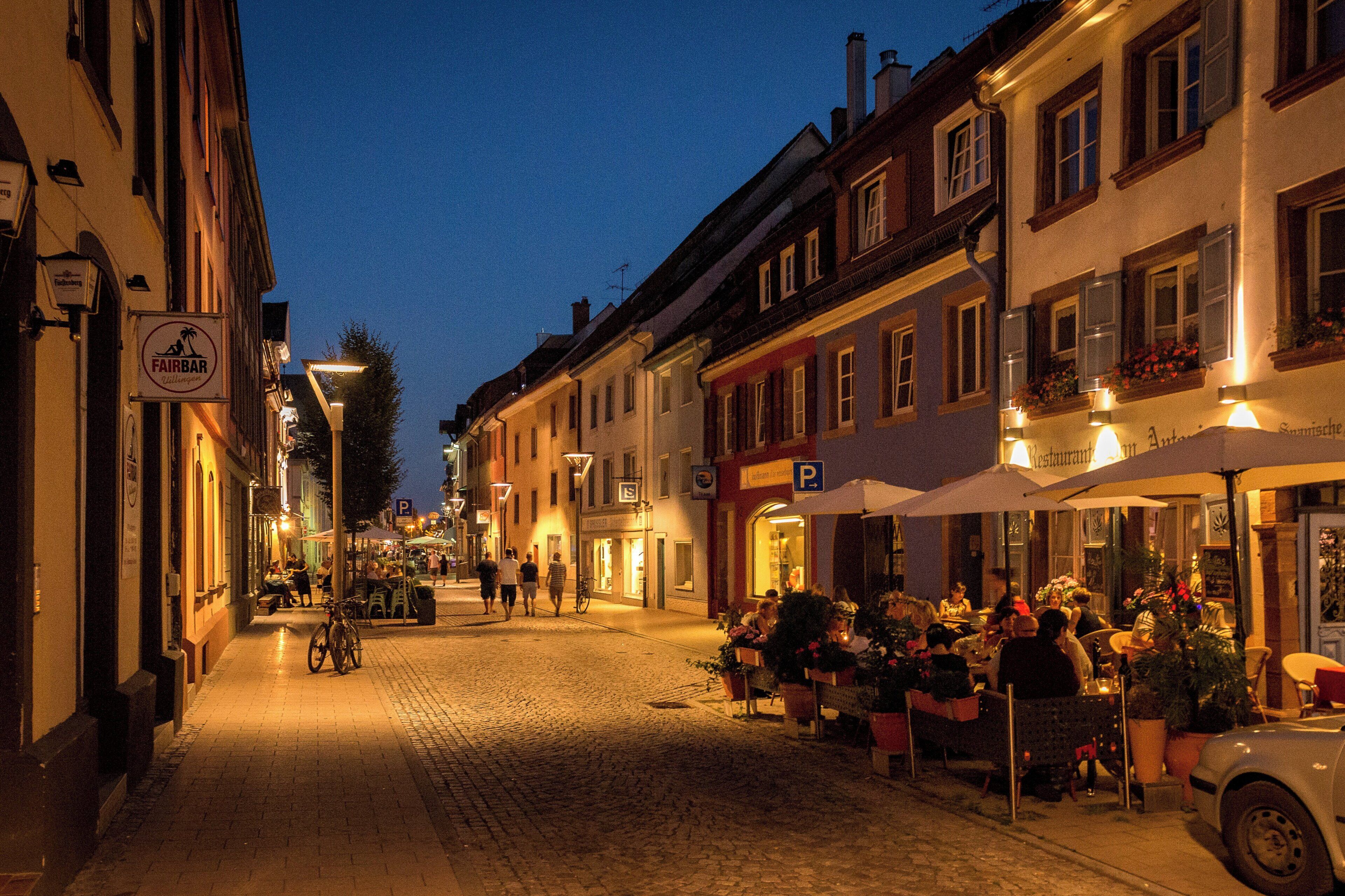 A cobblestone street lined with buildings, some with outdoor seating and umbrellas, indicating a dining area.