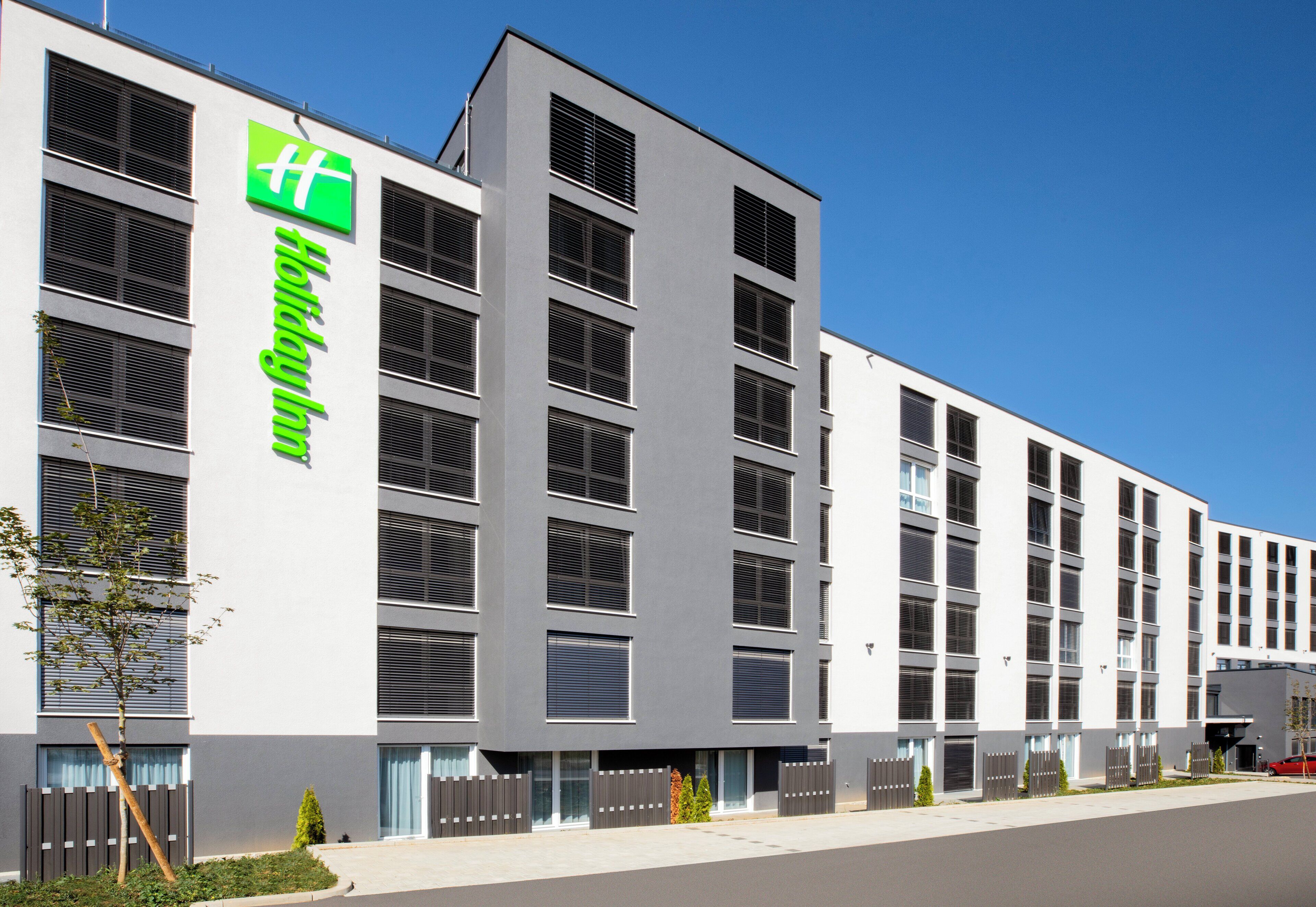 A modern hotel building with a prominent green logo, multiple windows, and a gated entrance.