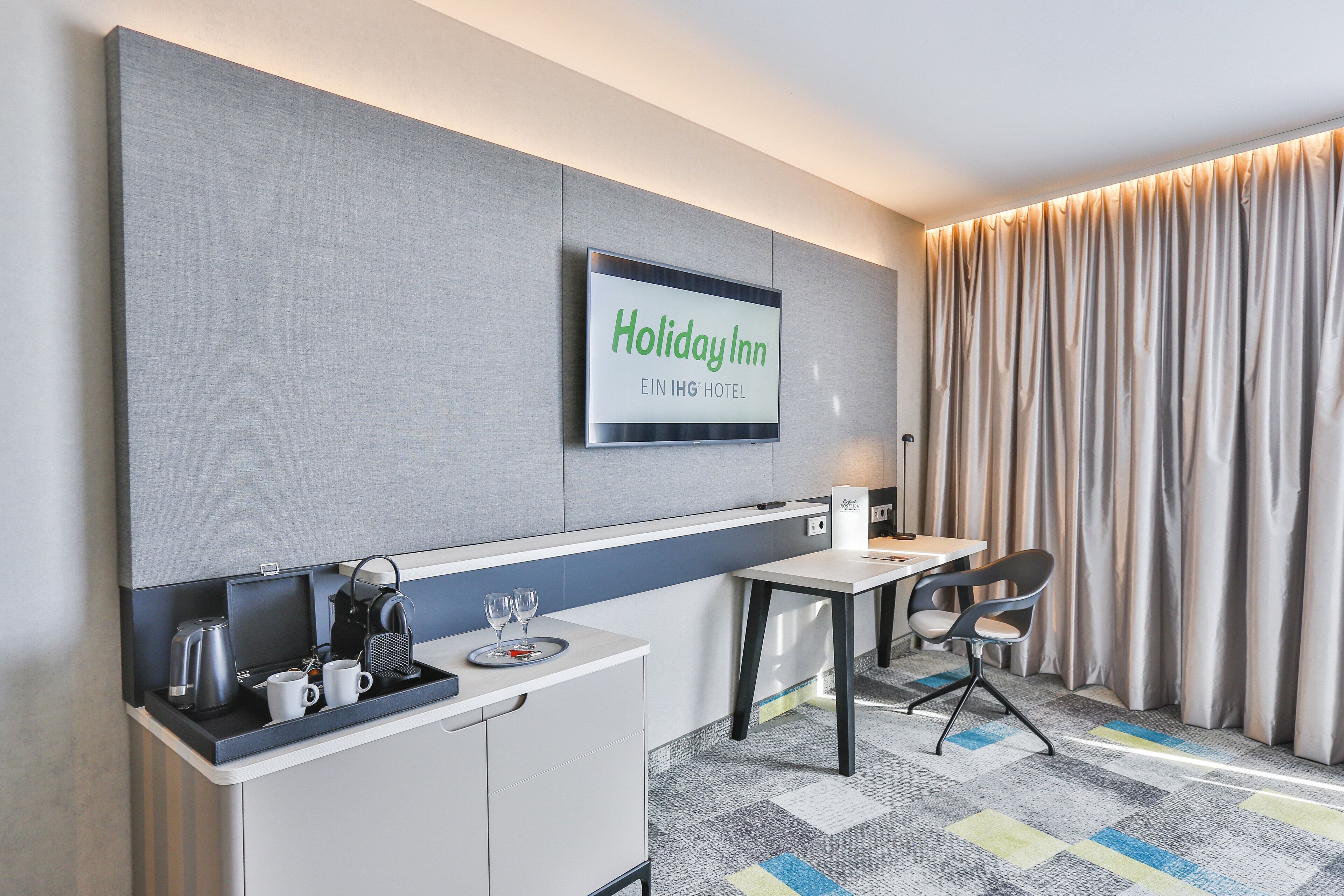A modern hotel room with a flat-screen TV displaying the Holiday Inn logo, a small kitchenette with a coffee maker and mugs, a dining area with a table and chairs, and a large window with curtains.