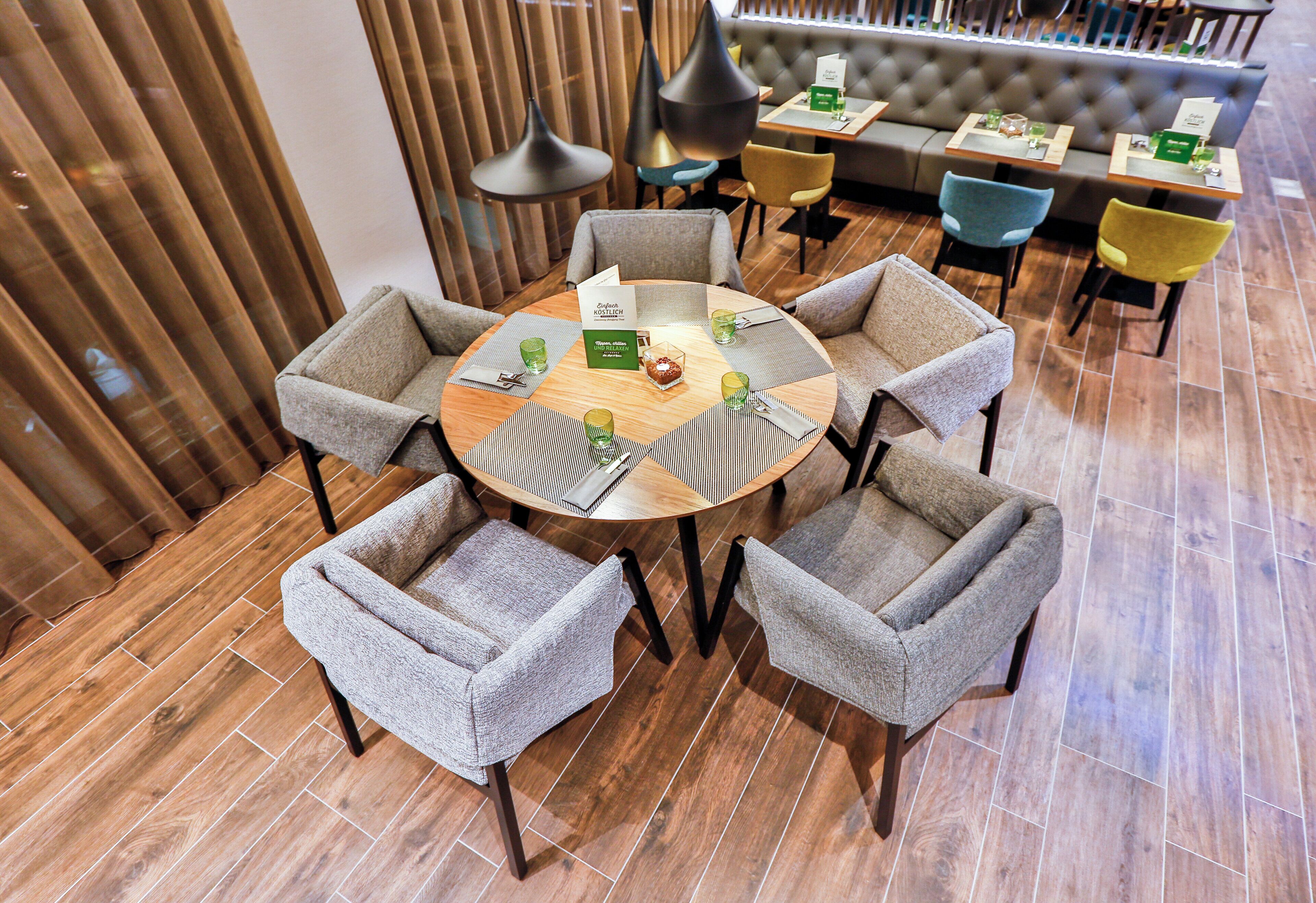 A round table with six grey upholstered chairs, surrounded by wooden flooring, in a modern interior with a sofa and a dining area in the background.