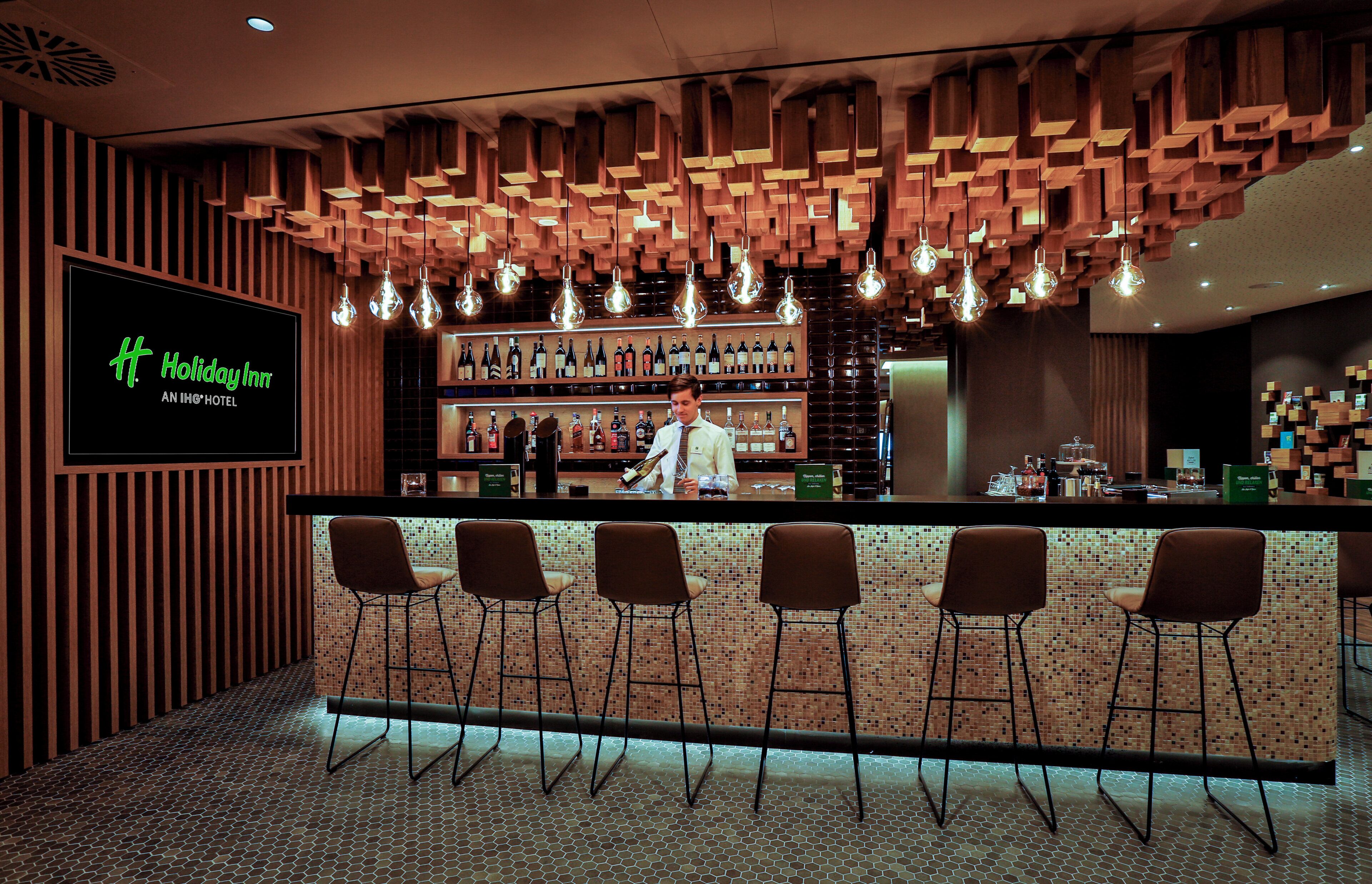 Zona de bar con techo de madera, lámparas colgantes y un camarero.