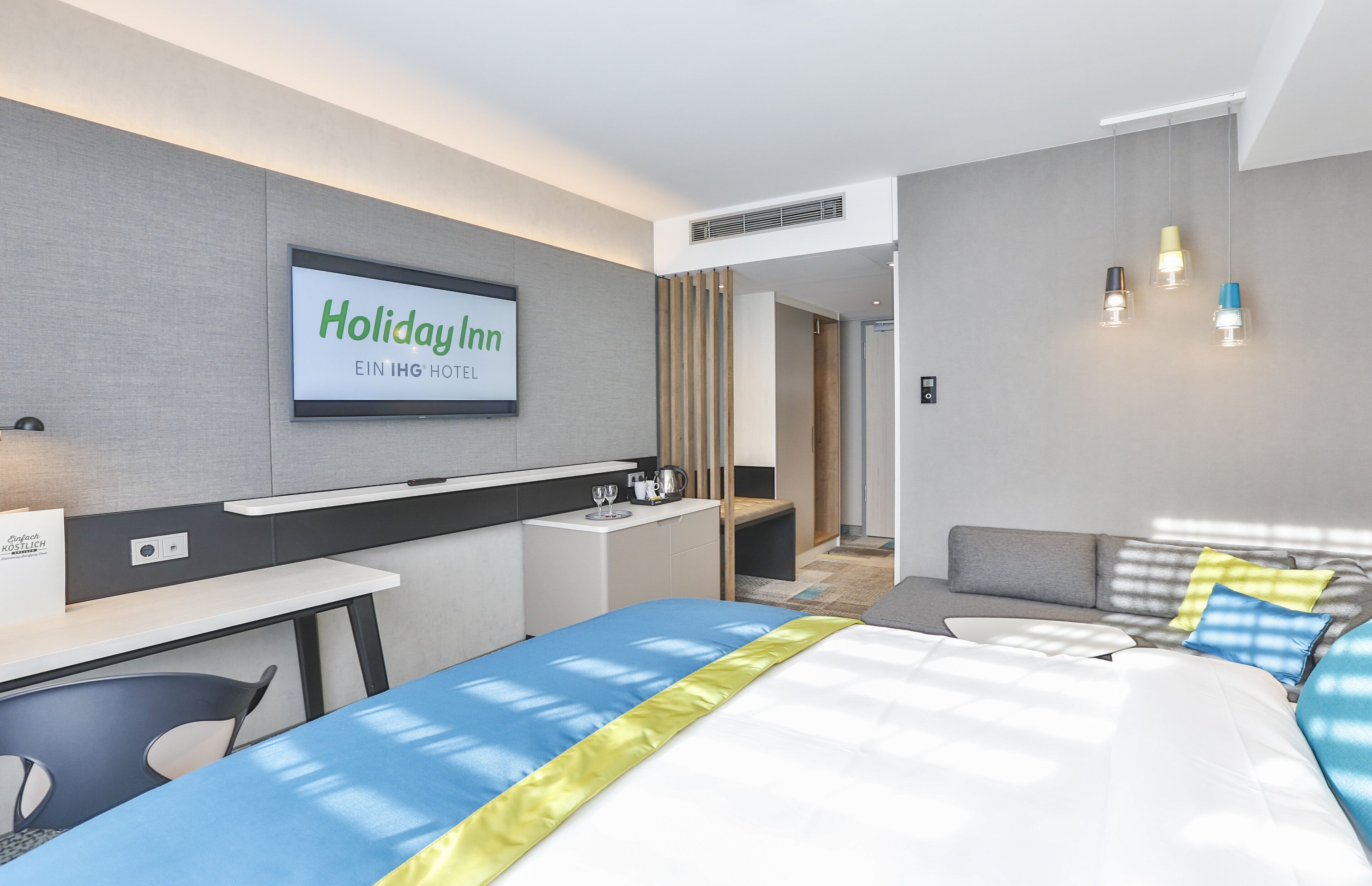 A modern hotel room with a bed, a desk, a chair, a TV displaying 'Holiday Inn EIN IHG HOTEL', and a sofa.