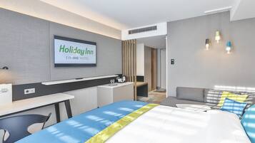 A modern hotel room with a bed, a desk, a chair, a TV displaying 'Holiday Inn EIN IHG HOTEL', and a sofa.