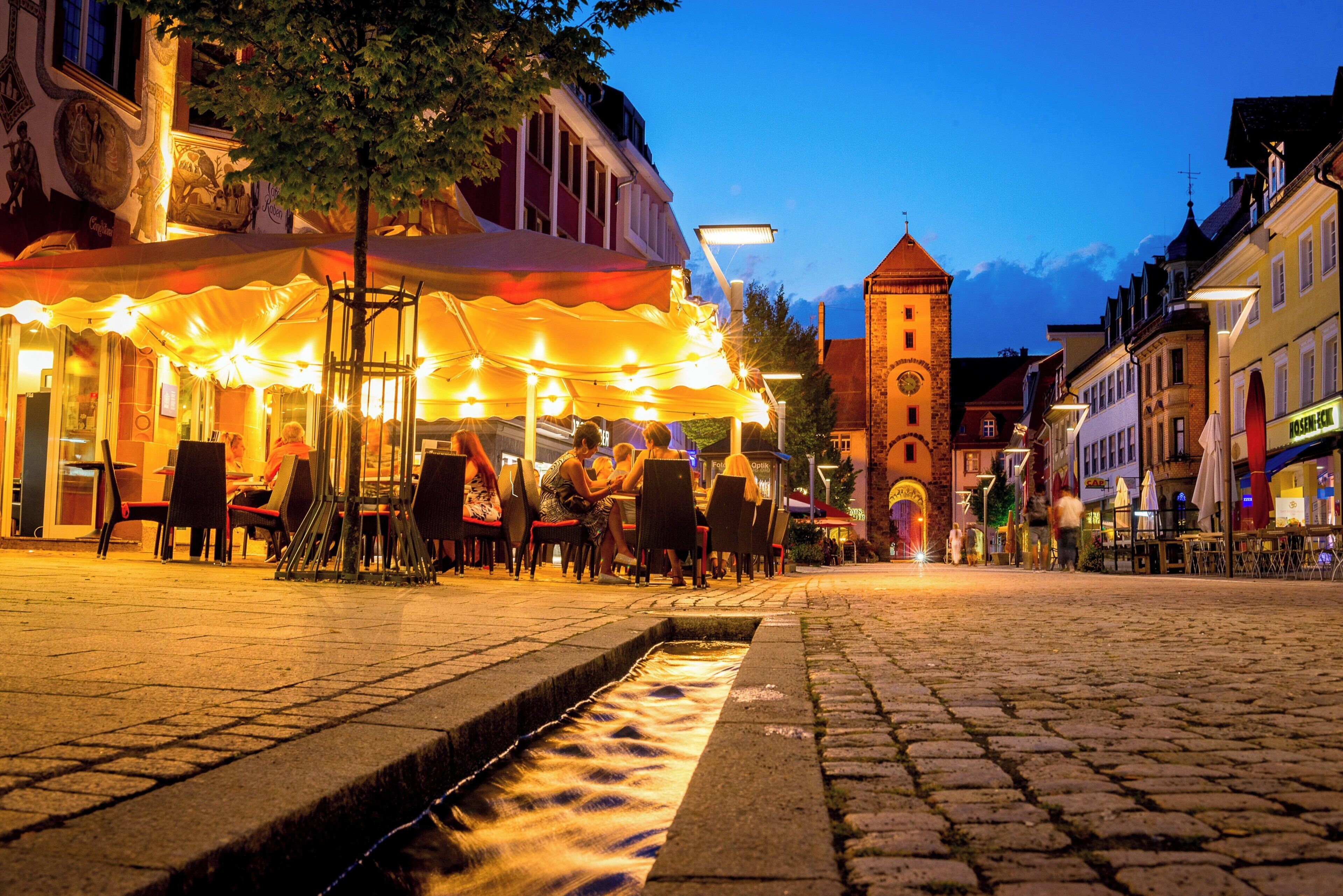 Uma rua de paralelepípedos com mesas ao ar livre em um café, um elemento aquático e edifícios históricos ao fundo.