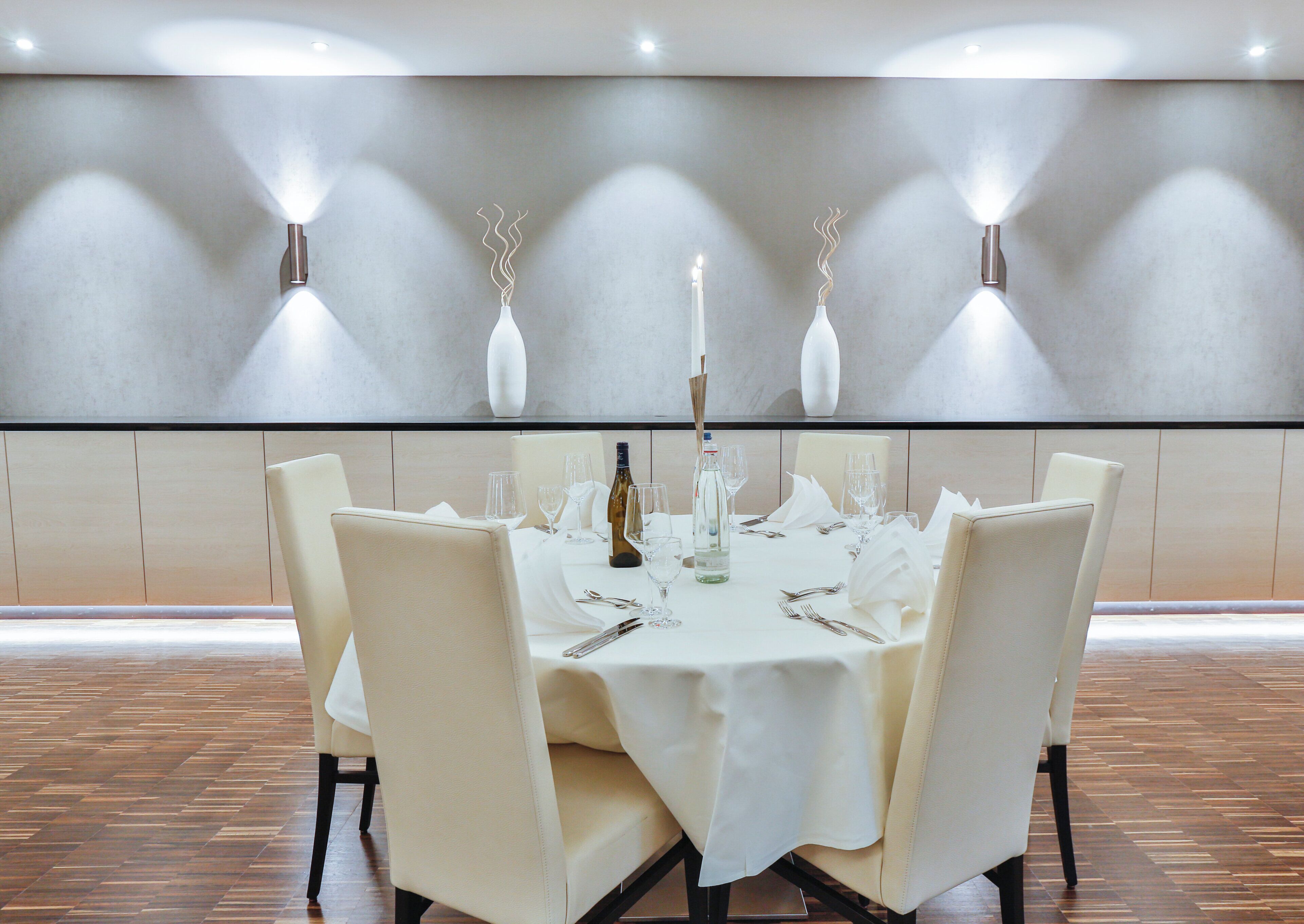 A dining table set for six with white tablecloth, candles, and wine bottles.