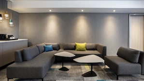 A modern living room with a grey sectional sofa, two small tables, and a kitchen area with a countertop and appliances. - Holiday Inn Villingen - Schwenningen by IHG (Villingen-Schwenningen)