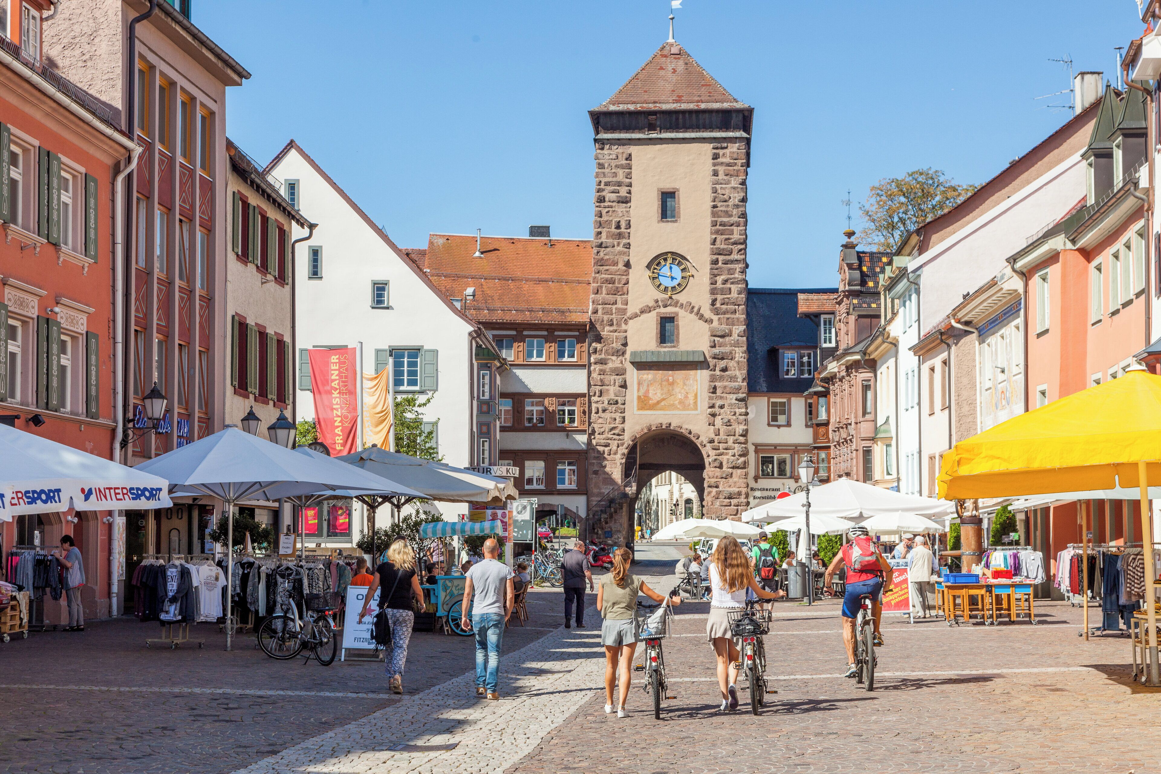 A cobblestone street with historic buildings, a clock tower, and outdoor market stalls.