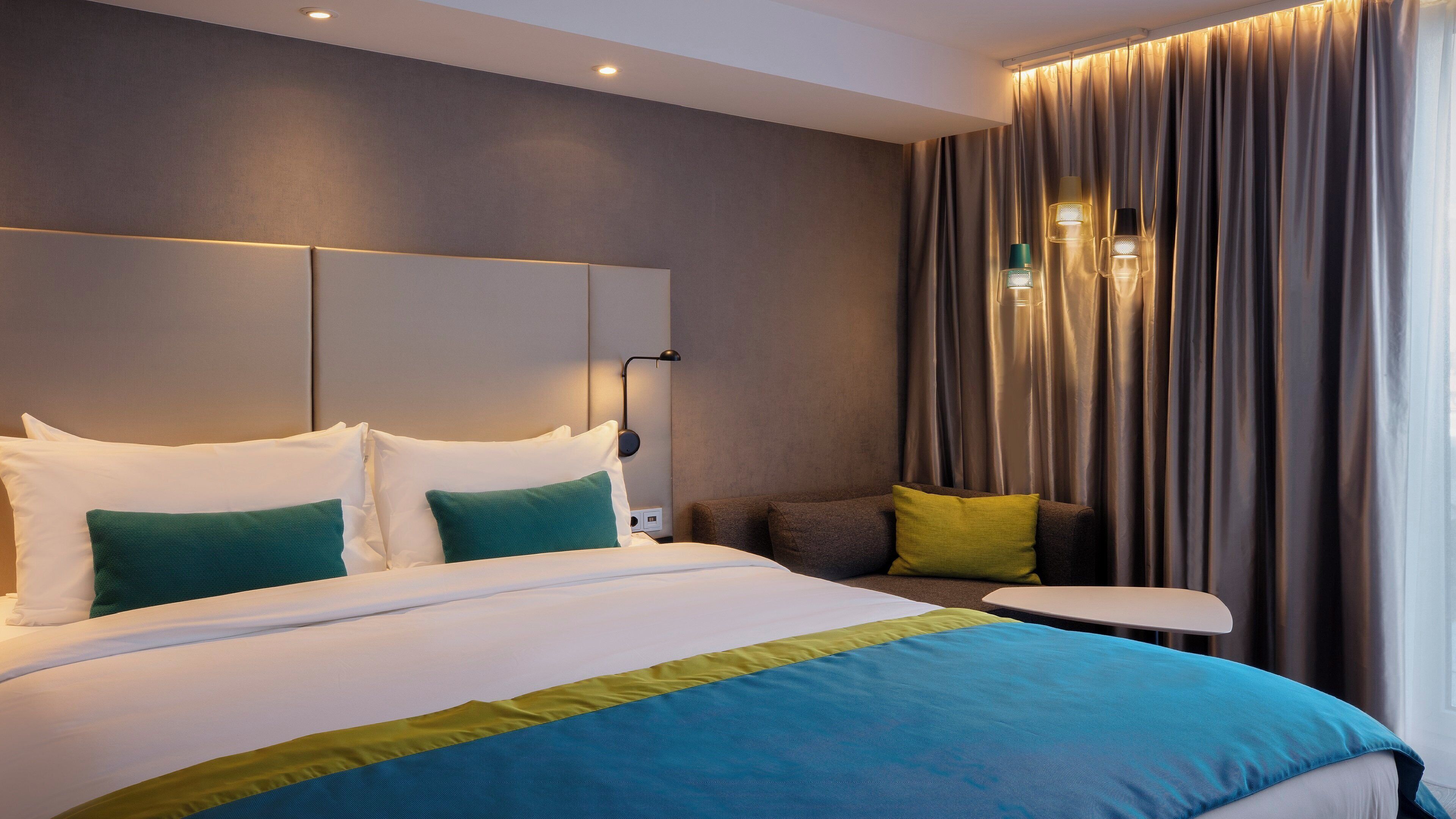 Une chambre d’hôtel moderne avec un grand lit, une table de chevet et une vue sur la ville, visible à travers les rideaux.