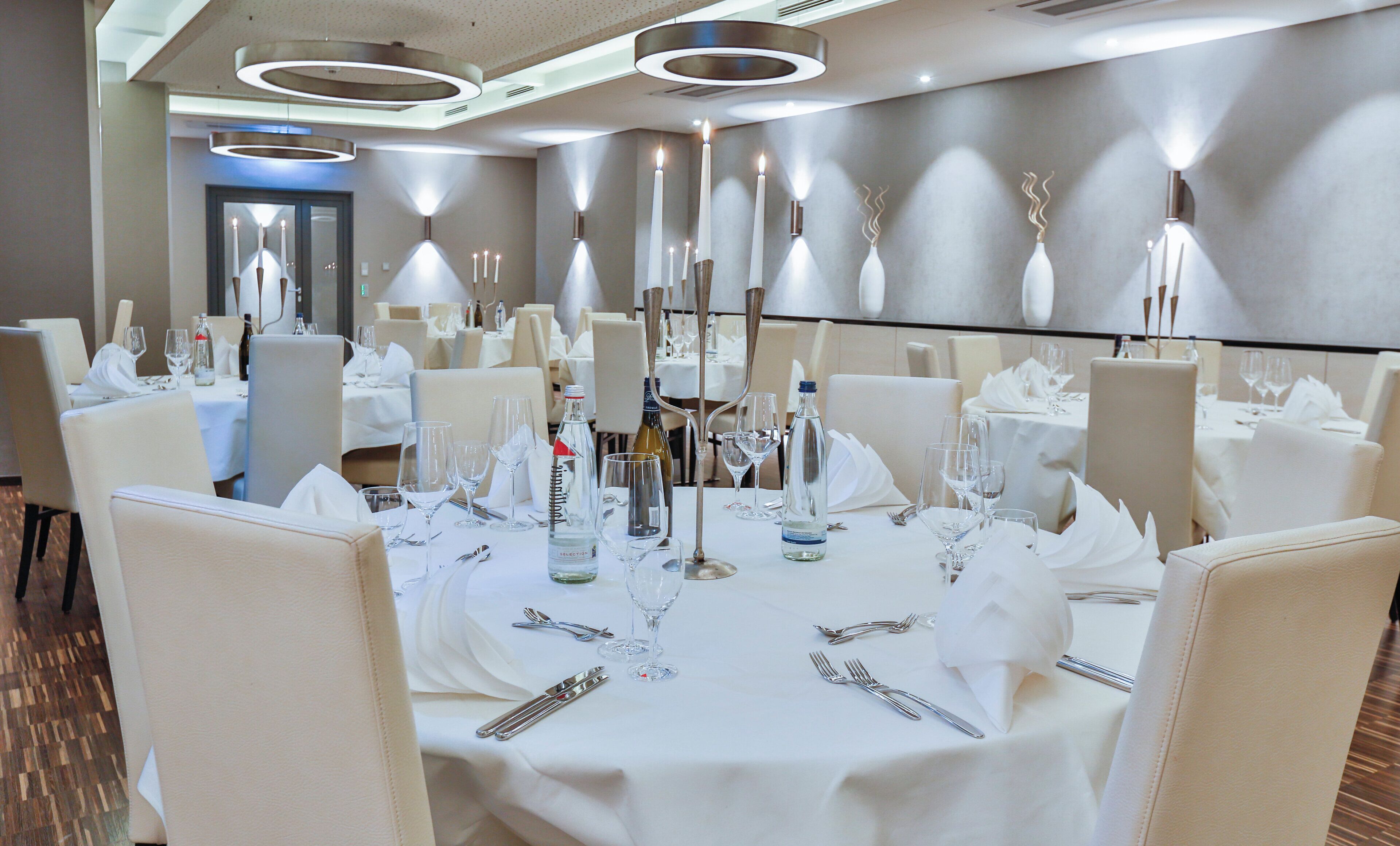 A hotel dining area with round tables set for a meal, each with cutlery, glasses, and a bottle of water.
