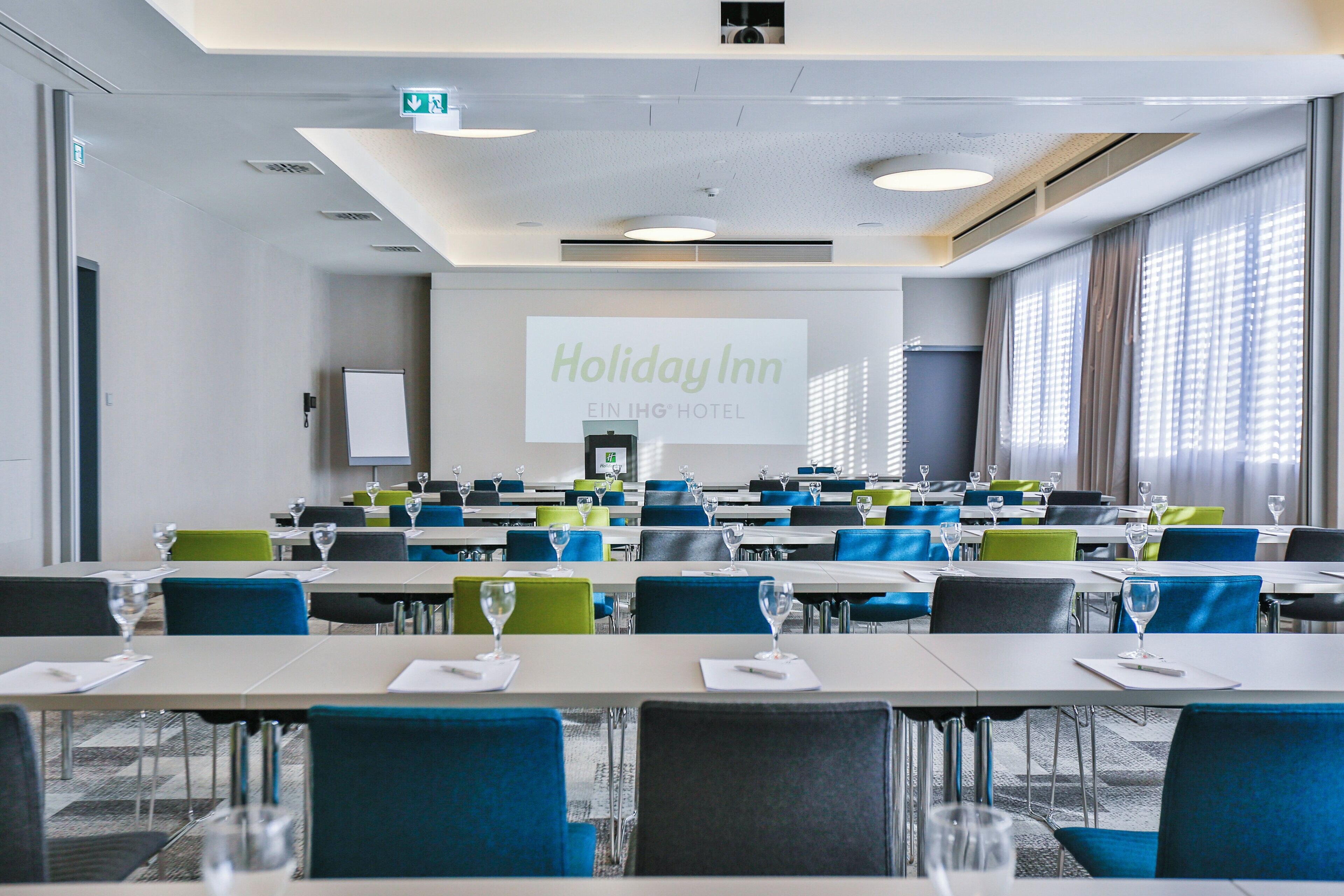 A conference room with tables and chairs, a projection screen displaying 'Holiday Inn EIN IHG HOTEL', and a large window with curtains.