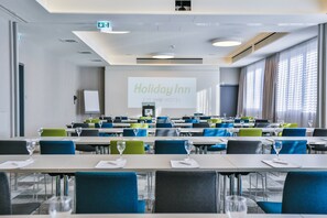 Una sala conferenze con tavoli e sedie, uno schermo di proiezione che mostra 'Holiday Inn EIN IHG HOTEL' e un'ampia finestra con tende.