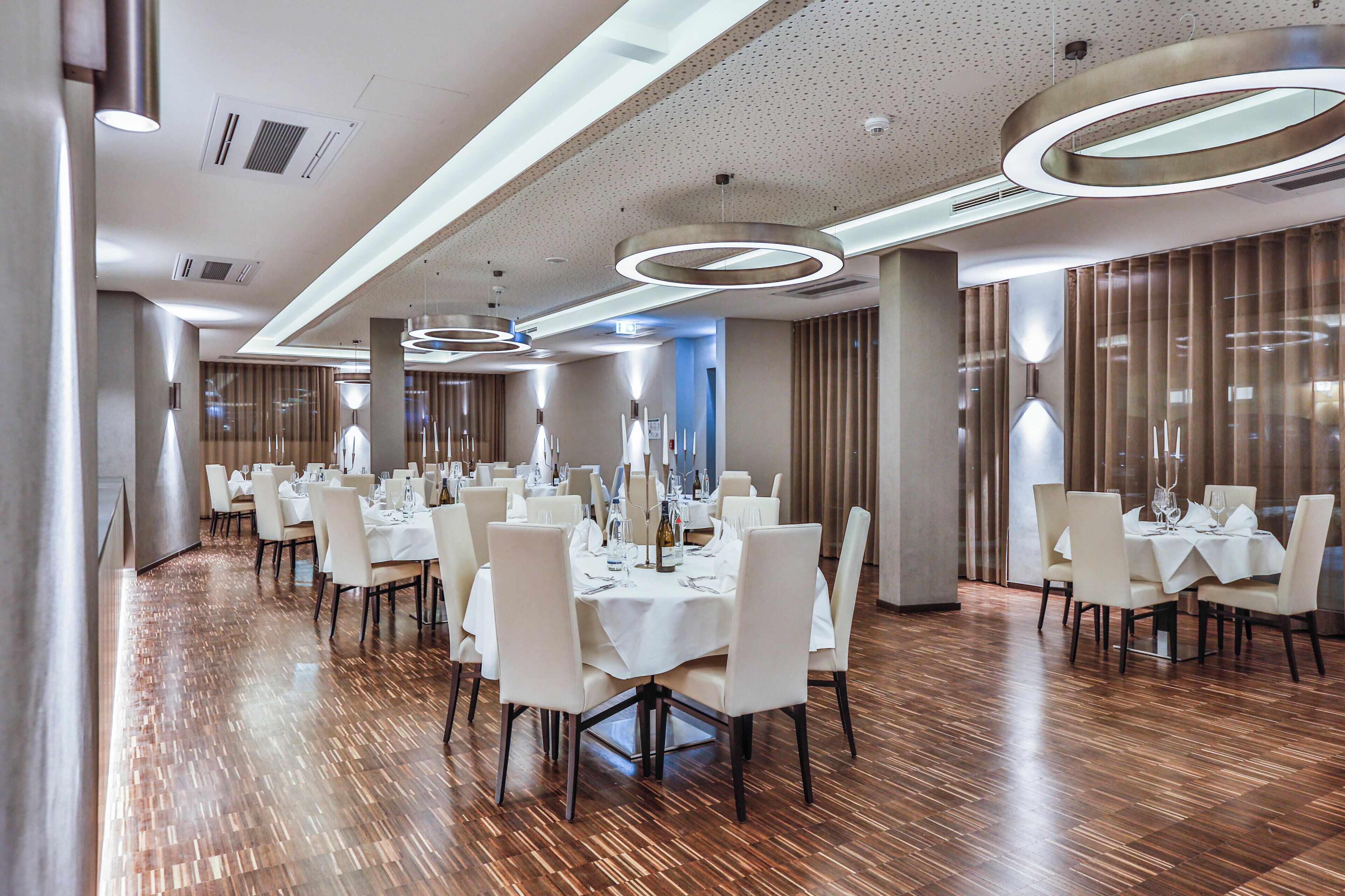 A spacious dining area with multiple tables and chairs, modern lighting fixtures, and a polished wooden floor.