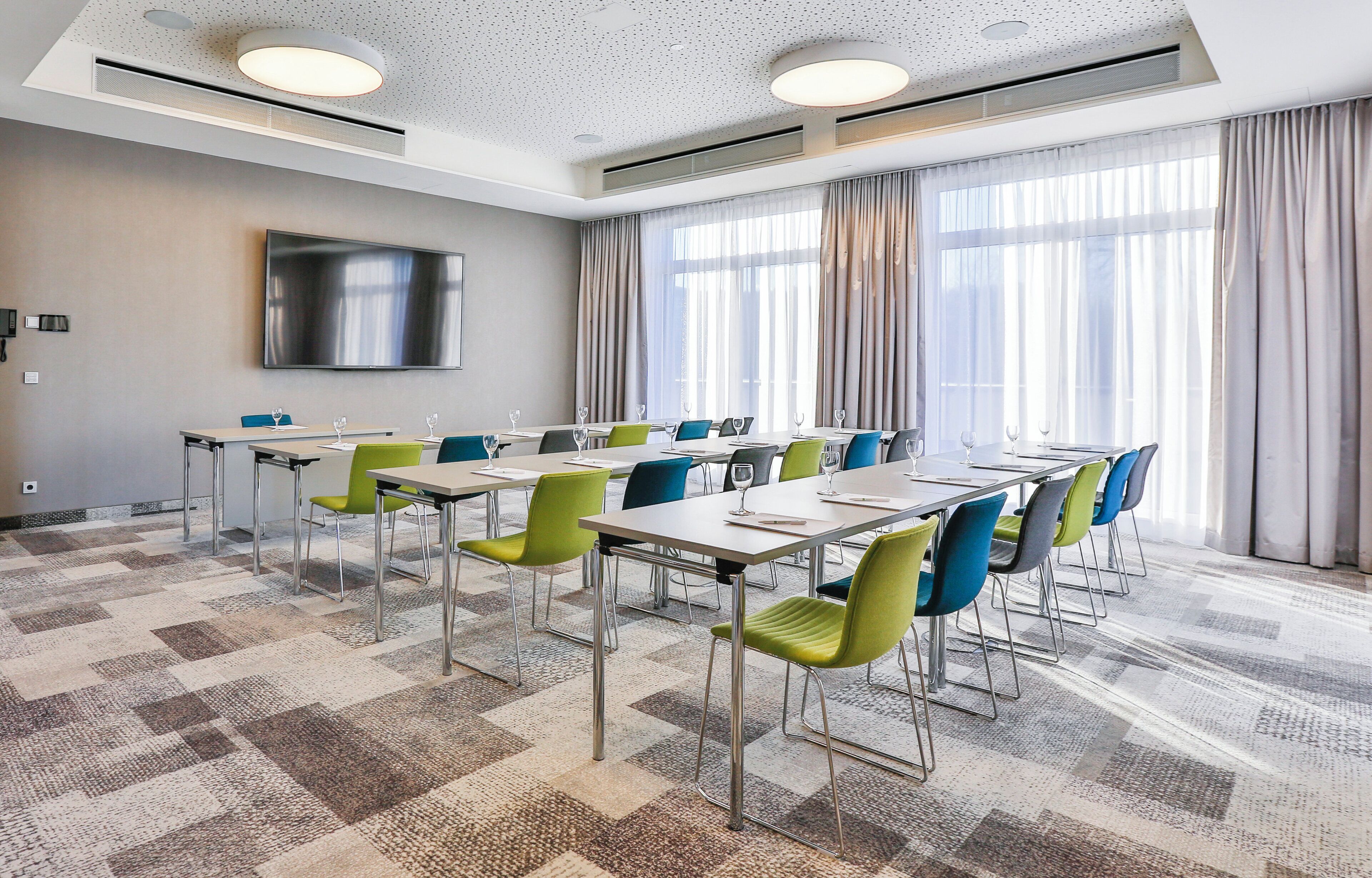 A conference room with tables, chairs, a flat-screen TV, and large windows with curtains.