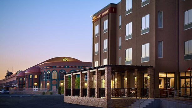 A hotel with a modern design, featuring a prominent sun emblem on the facade, a covered outdoor area with stone pillars, and a large dome structure in the background. - The Hotel at Sunland Park Casino El Paso, an Ascend Collection Hotel (Sunland Park)