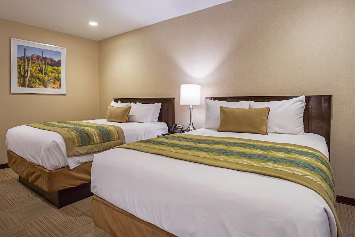 A hotel room with two beds, beige walls, a framed picture of a desert landscape, and a bedside lamp.